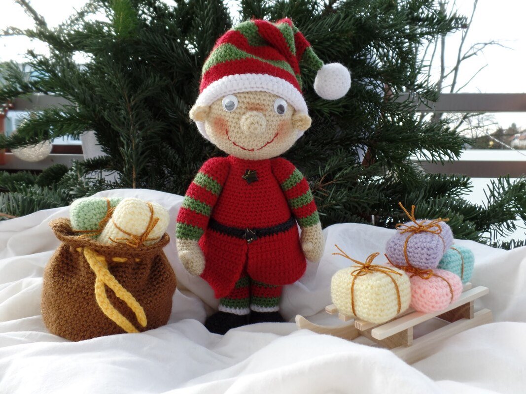 Gehäkelter Weihnachtself in rot-grün mit Zipfelmütze neben Geschenkesack und Schlitten