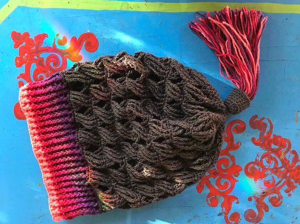 Dunkelgraue gehäkelte Damenmütze mit rosa-lilafarbenem Bund und mehrfarbiger Quaste auf blauem Hintergrund