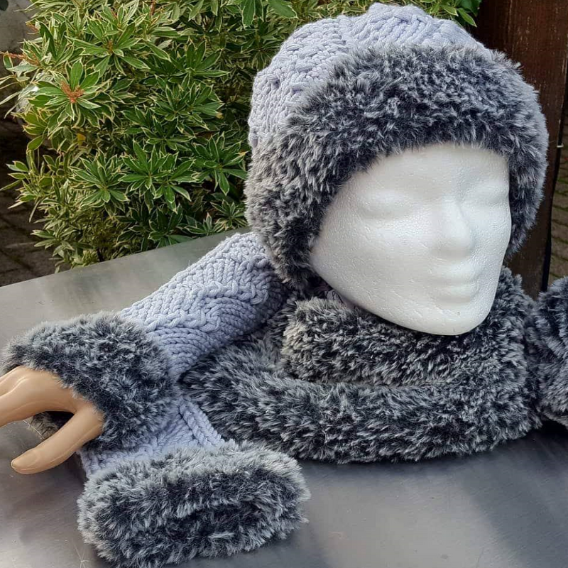 Strickset aus Mütze, Schal und Armstulpen in blaugrauem Zopfmuster mit dunklem Kunstfellbesatz, präsentiert auf einer Schaufensterpuppe.
