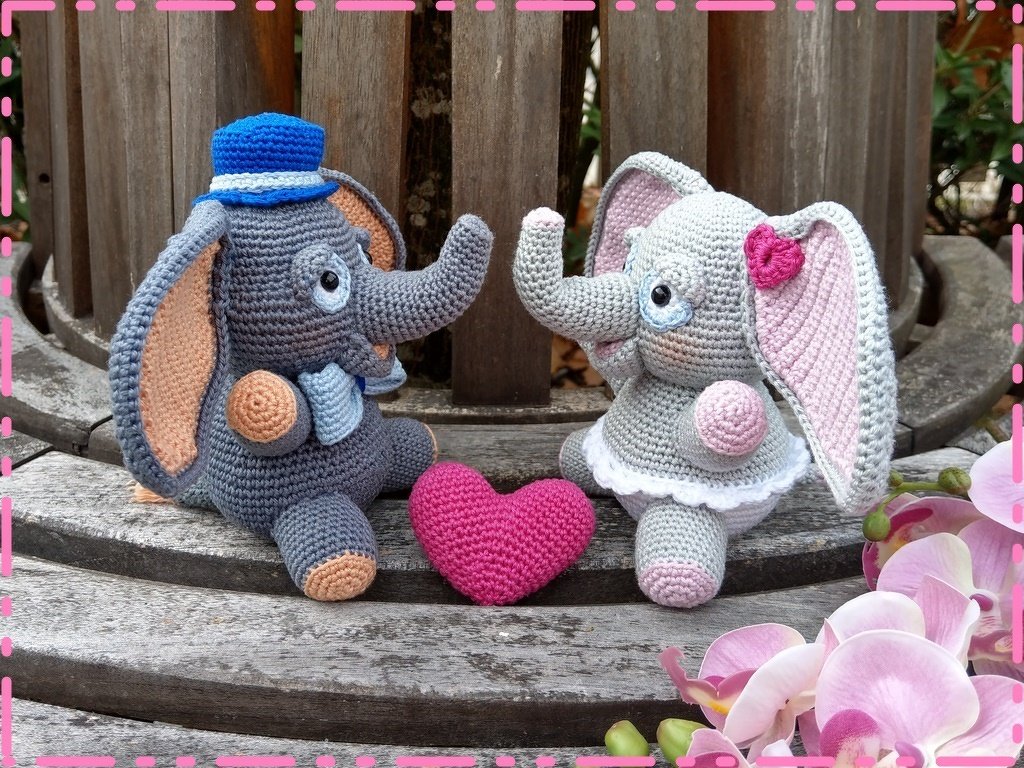 Zwei gehäkelte Elefanten-Amigurumis mit blauem Hut und pinker Schleife neben einem pinken Häkelherz auf Holzbank.