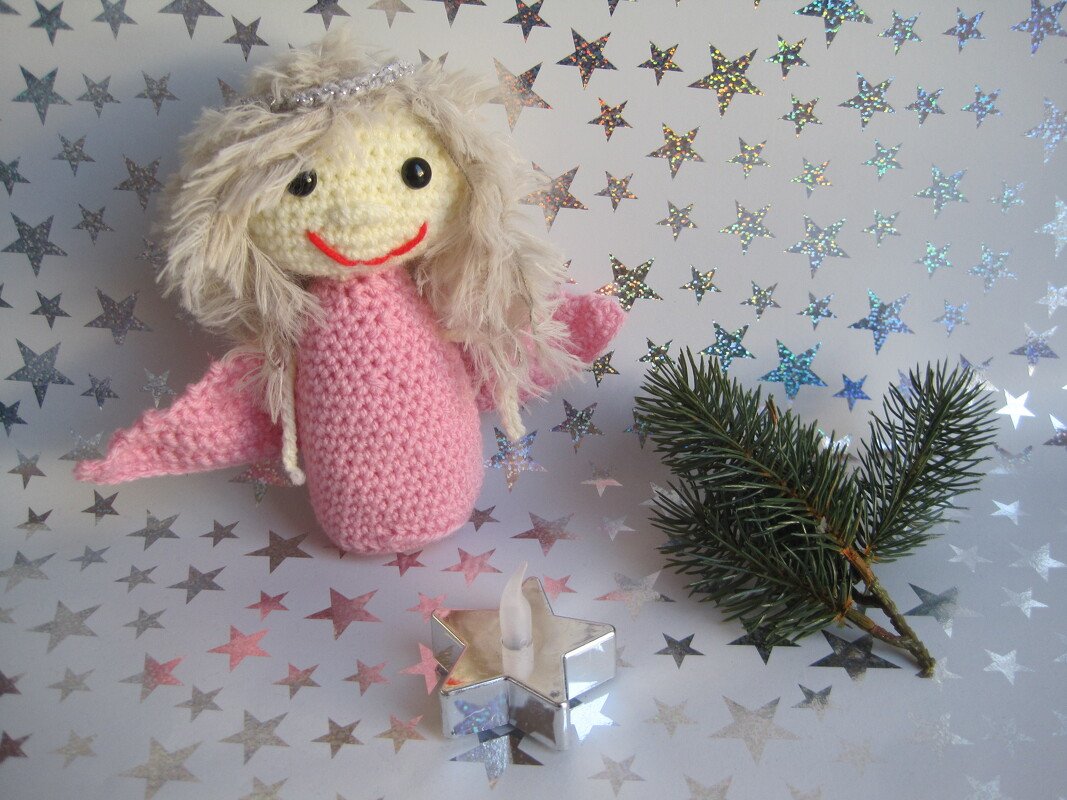 Rosa gehäkeltes Engelchen mit hellen flauschigen Haaren vor silbernem Sternenmuster, neben einem Tannenzweig