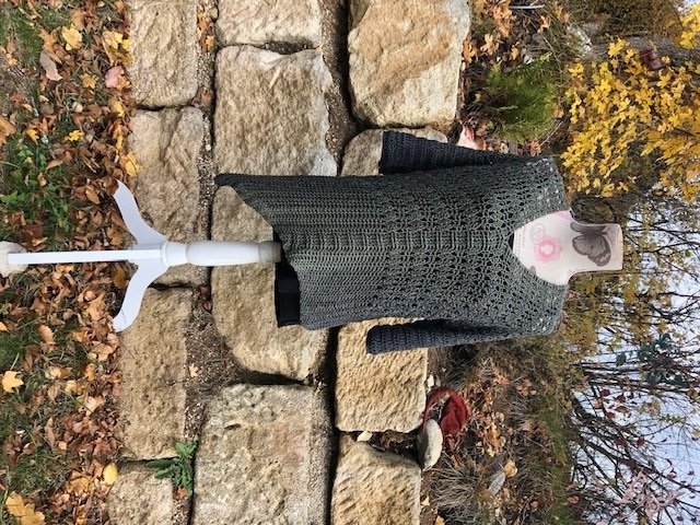 Dunkelgrünes Häkeloberteil mit langen Ärmeln auf Schaufensterpuppe vor Steinen