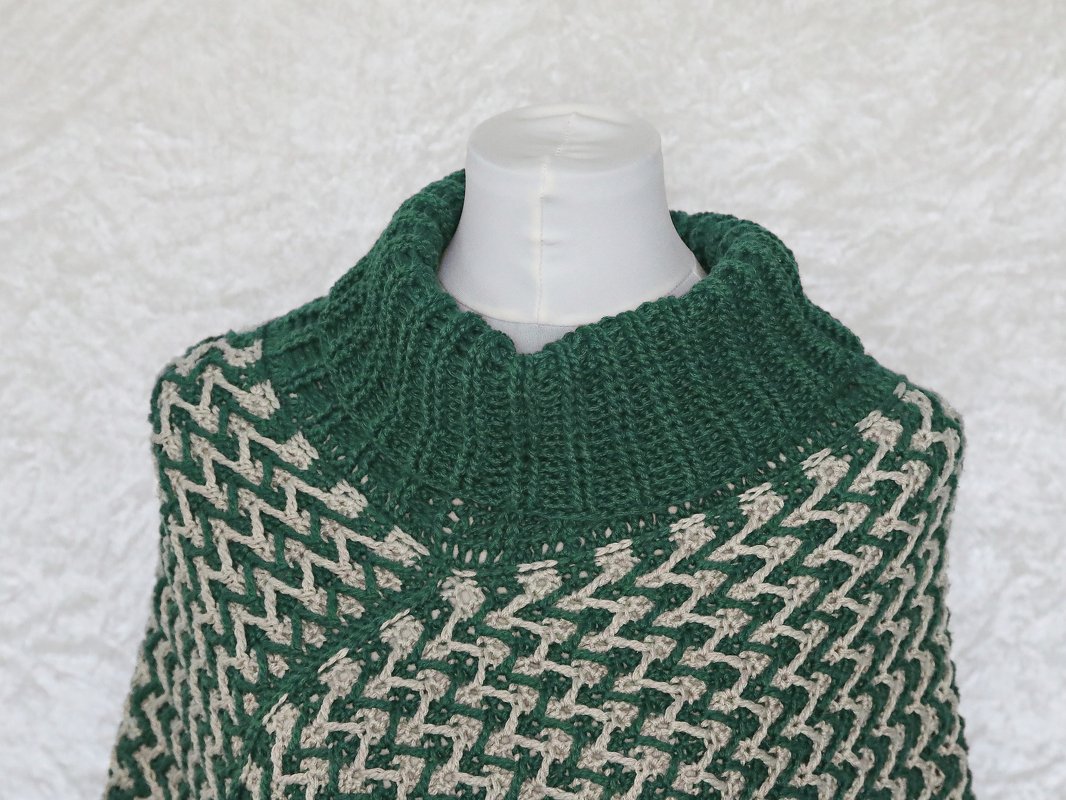 Gehäkelter Damen-Poncho in Dunkelgrün und Beige mit grafischem Zickzackmuster und hohem Rollkragen auf einer Schneiderpuppe.