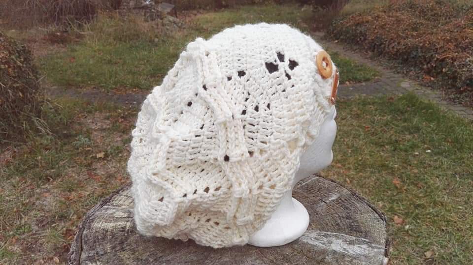 Cremeweiße gehäkelte Beanie mit Relief- und Lochmuster und zwei Holzknöpfen auf einer Büste im Freien.