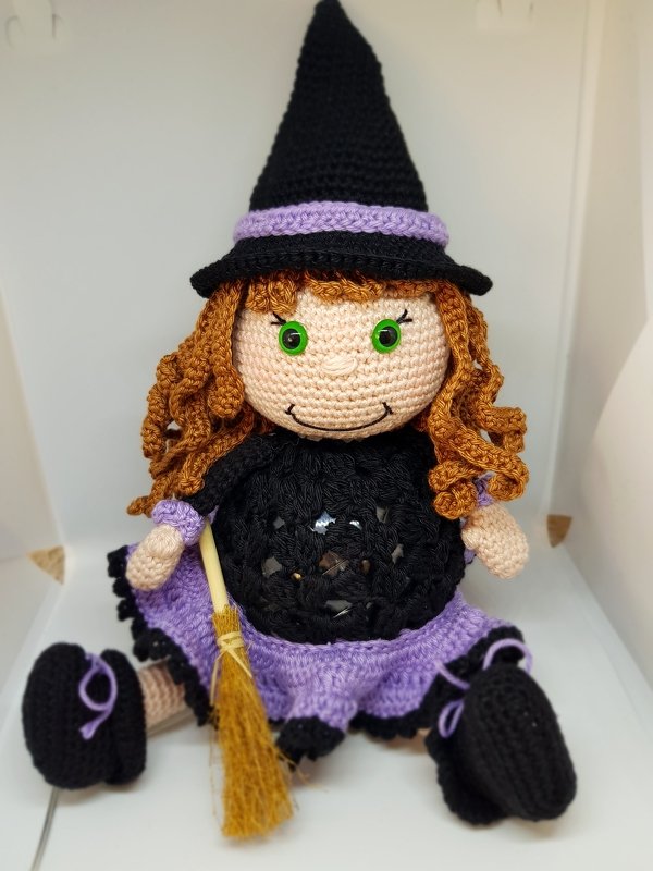 Gehäkelte Hexenpuppe mit rötlich-braunen Locken, grünem Augenpaar, schwarzem Hut, lila Rock und kleinem Besen