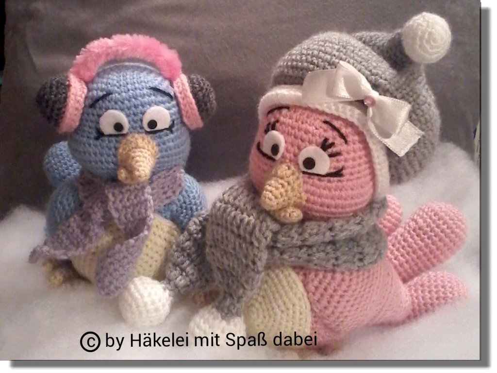 Zwei gehäkelte Vögel (blau mit pinken Ohrenschützern, rosa mit grauer Mütze) auf weißem Untergrund.