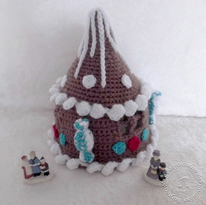 Gehäkeltes braunes Lebkuchenhaus mit weißer Dach- und Randverzierung und bunten Akzenten, dazu zwei kleine Figuren