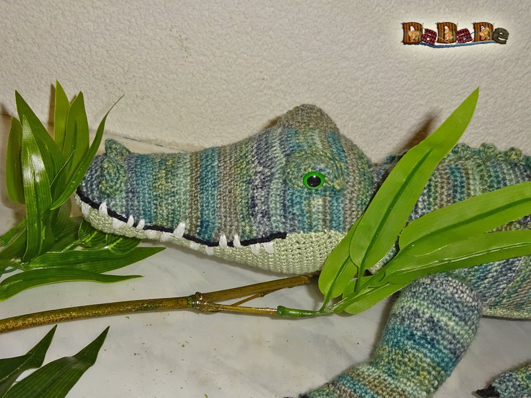 Gehäkeltes Amigurumi-Krokodil aus meliertem Garn in Grün- und Blautönen mit grünen Augen und angedeuteten weißen Zähnen, dekoriert mit grünen Blättern.
