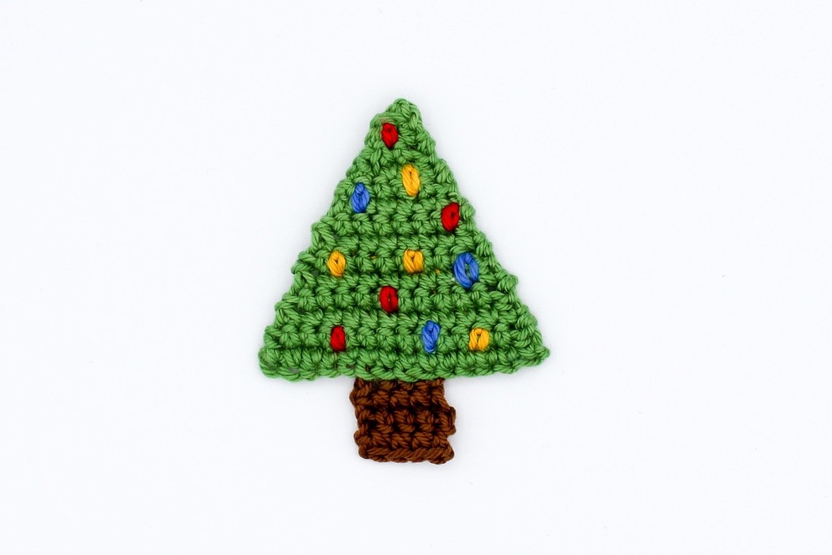 Gehäkelte grüne Weihnachtsbaum-Applikation mit bunten Zierpunkten und braunem Stamm vor weißem Hintergrund