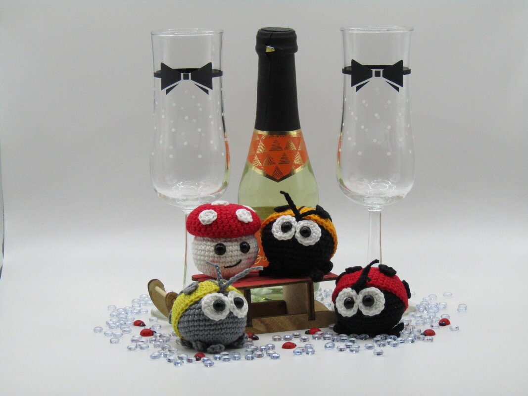 Mehrere gehäkelte Amigurumi-Glücksbringer (Marienkäfer und Pilz) vor einer Sektflasche und zwei Sektgläsern auf weißem Hintergrund