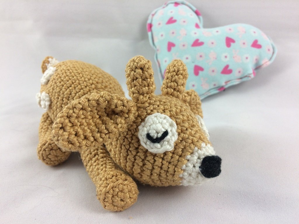 Gehäkeltes liegendes Rehkitz-Amigurumi aus braunem Garn vor blau-rosa Herzkissen