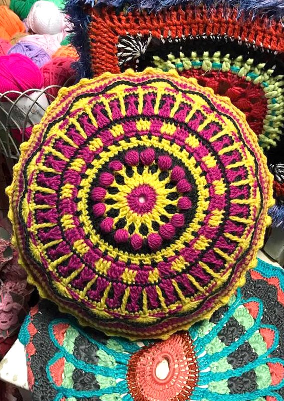 Detailaufnahme eines runden gehäkelten Kissens mit Noppenstich, gelben und pinken Ringen, weiterer Häkelware und Garn im Hintergrund