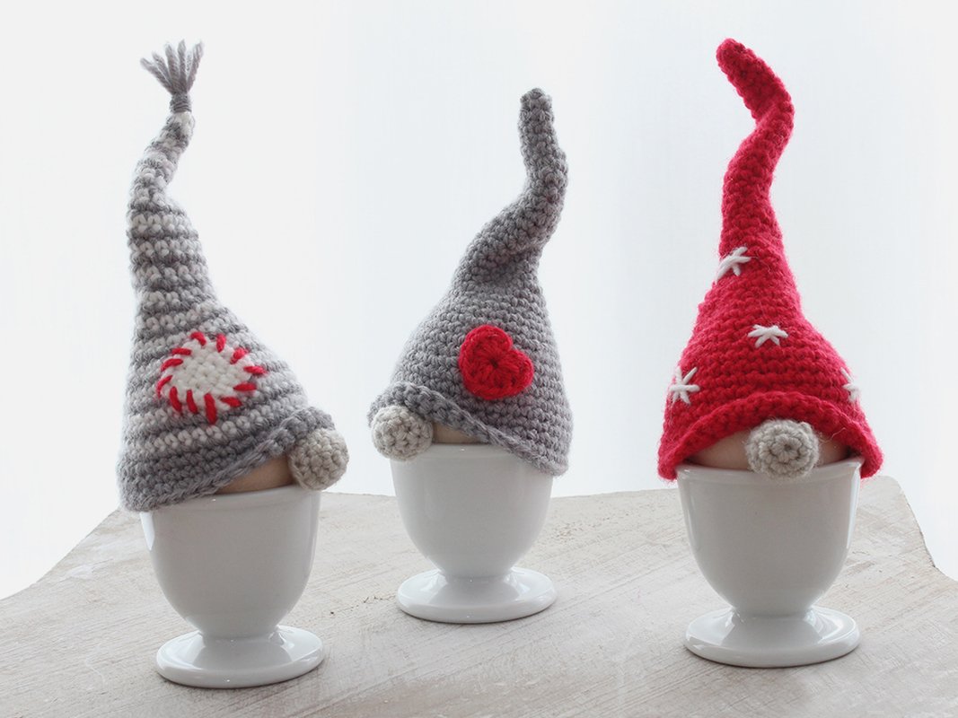 Three crochet gnome egg cosies on eggs in white egg cups, two grey and one red