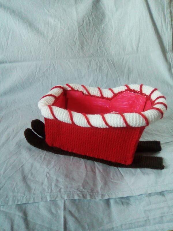 Gehäkelter roter Weihnachtsschlitten mit weißem, gedrehtem Rand und braunen Kufen auf hellem Stoff