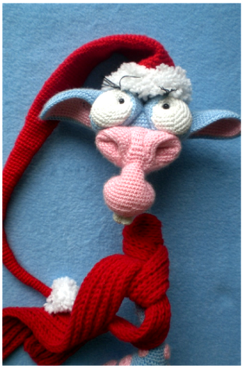 Gehäkelter Fantasie-Tierkopf in Blau und Rosa mit großen weißen Augen, roter Zipfelmütze und rotem Schal auf blauem Hintergrund