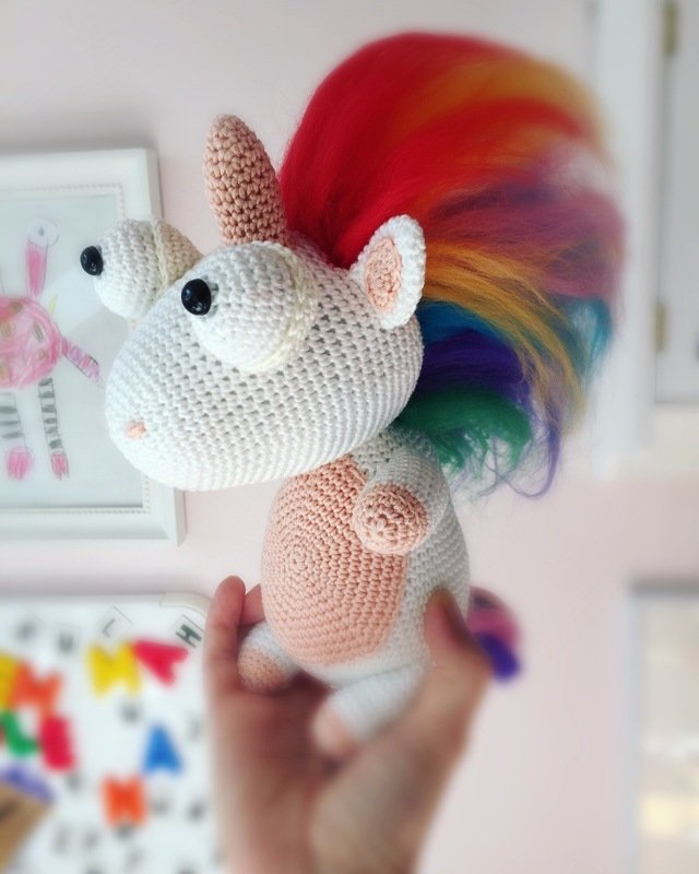Nahaufnahme eines gehäkelten Einhorn-Amigurumi mit weißem Körper, rosa Bauch und bunter Regenbogenmähne, in einer Hand gehalten.