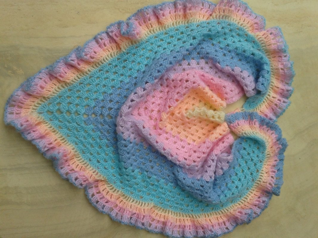 Gehäkeltes Dreieckstuch in Pastellfarben mit Granny‑Square‑Muster und Rüschenrand auf hellem Hintergrund.