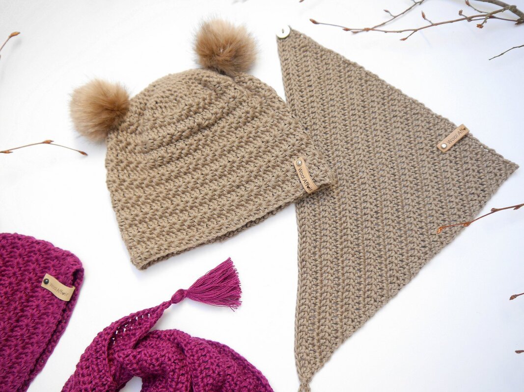 Beige crocheted baby beanie with two faux-fur pom-poms next to a beige triangular crochet bandana with a button; magenta crochet pieces partially visible