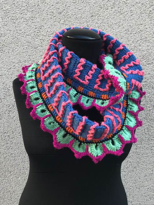 Bunter gehäkelter Loop-Schal mit Wellenmuster und Muschelrand auf schwarzer Büste.