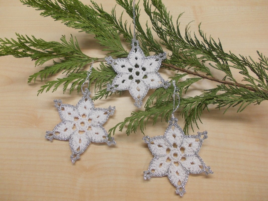 Drei gehäkelte weiße Schneeflocken-Anhänger mit silbernem Rand auf hellem Holz neben einem Tannenzweig.