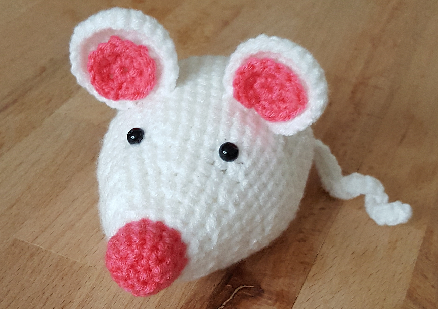 Gehäkelte weiße Amigurumi‑Maus mit rosa Nase, rosa Innenohren, schwarzen Augen und spiraligem Schwanz auf Holzfläche