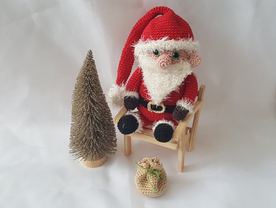 Gehäkelter Weihnachtsmann-Amigurumi in rotem Kostüm auf kleinem Holzstuhl, daneben goldene Tanne und gehäkelter Sack