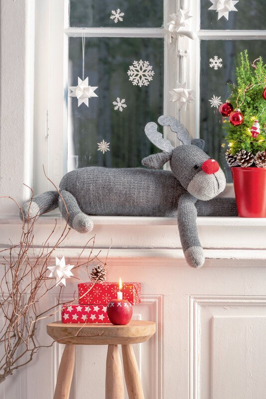 Gestricktes graues Rentier mit roter Nase auf der Fensterbank, umgeben von Papiersternen, Schneeflockenstickern und einem Mini-Weihnachtsbaum.