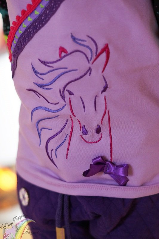 Stickerei eines stilisierten Pferdekopfes mit violetter, rosa und blauer Mähne auf hellviolettem Stoff, verziert mit einer kleinen violetten Schleife.