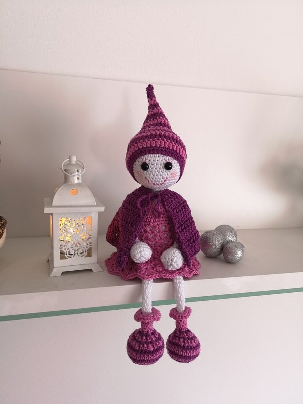 Handgehäkelte Amigurumi-Puppe in Lila und Rosa mit gestreifter Zipfelmütze auf weißem Regal neben einer Laterne