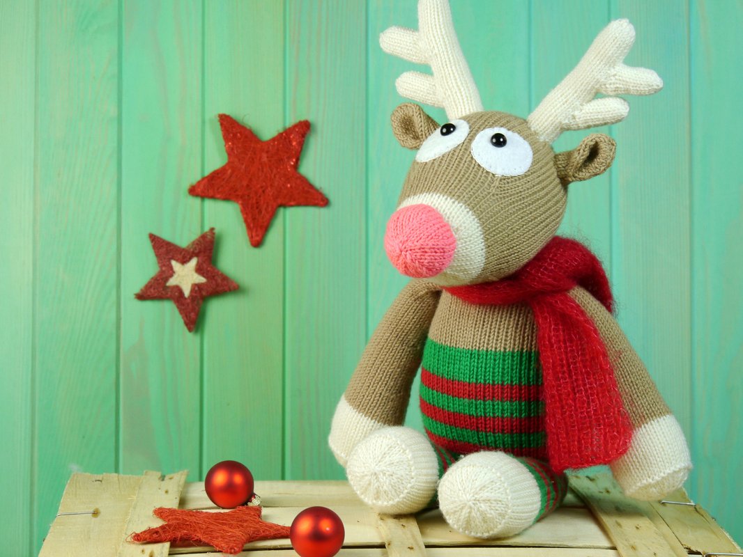 Knitted reindeer plush toy with red scarf, striped body, red stars, and baubles on a wooden surface against a green background.