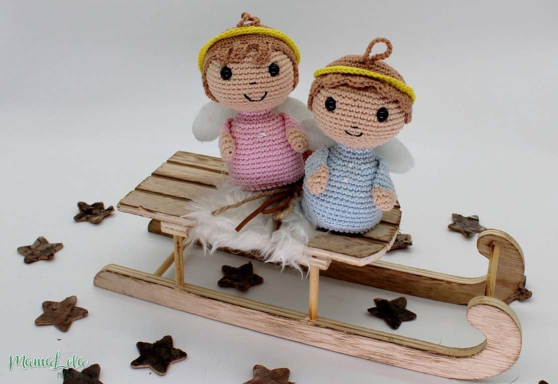 Zwei gehäkelte Engelchen mit weißen Flügeln in Rosa und Blau sitzen auf einem Holzschlitten, umgeben von Holzsternen