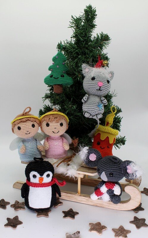 Verschiedene gehäkelte Anhänger (Engel, Pinguin, Katze, Koala, Tanne, Kerze) mit Schlaufen, arrangiert um einen Mini‑Schlitten vor einem kleinen Tannenbaum