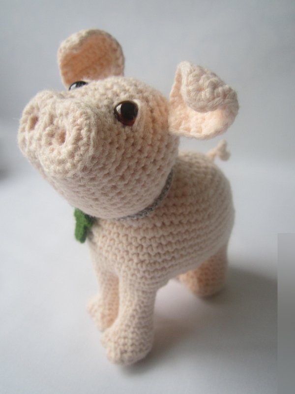 Helles gehäkeltes Amigurumi-Schwein mit aufgerichtetem Kopf, modellierter Schnauze und kleinem grünen Kleeblatt am Hals auf weißem Hintergrund.