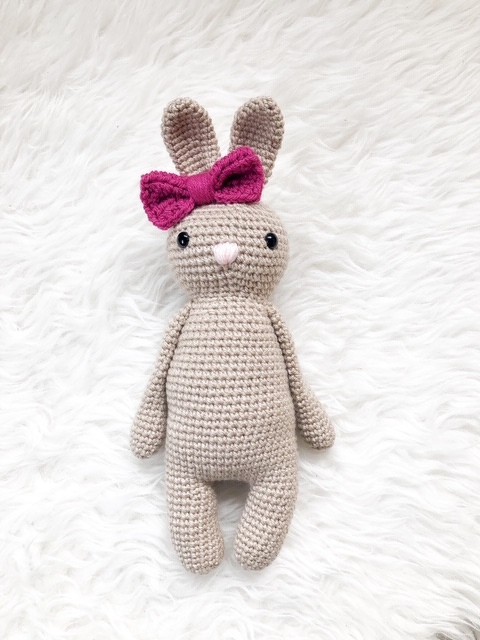 Gehäkelter beiger Amigurumi-Hase mit pinker Schleife auf weißem Plüsch