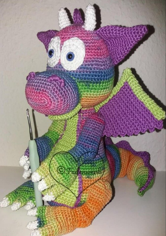 Gehäkelter Regenbogen-Drache mit großen weißen Augen, lila Flügeln und sichtbarem Häkelhaken