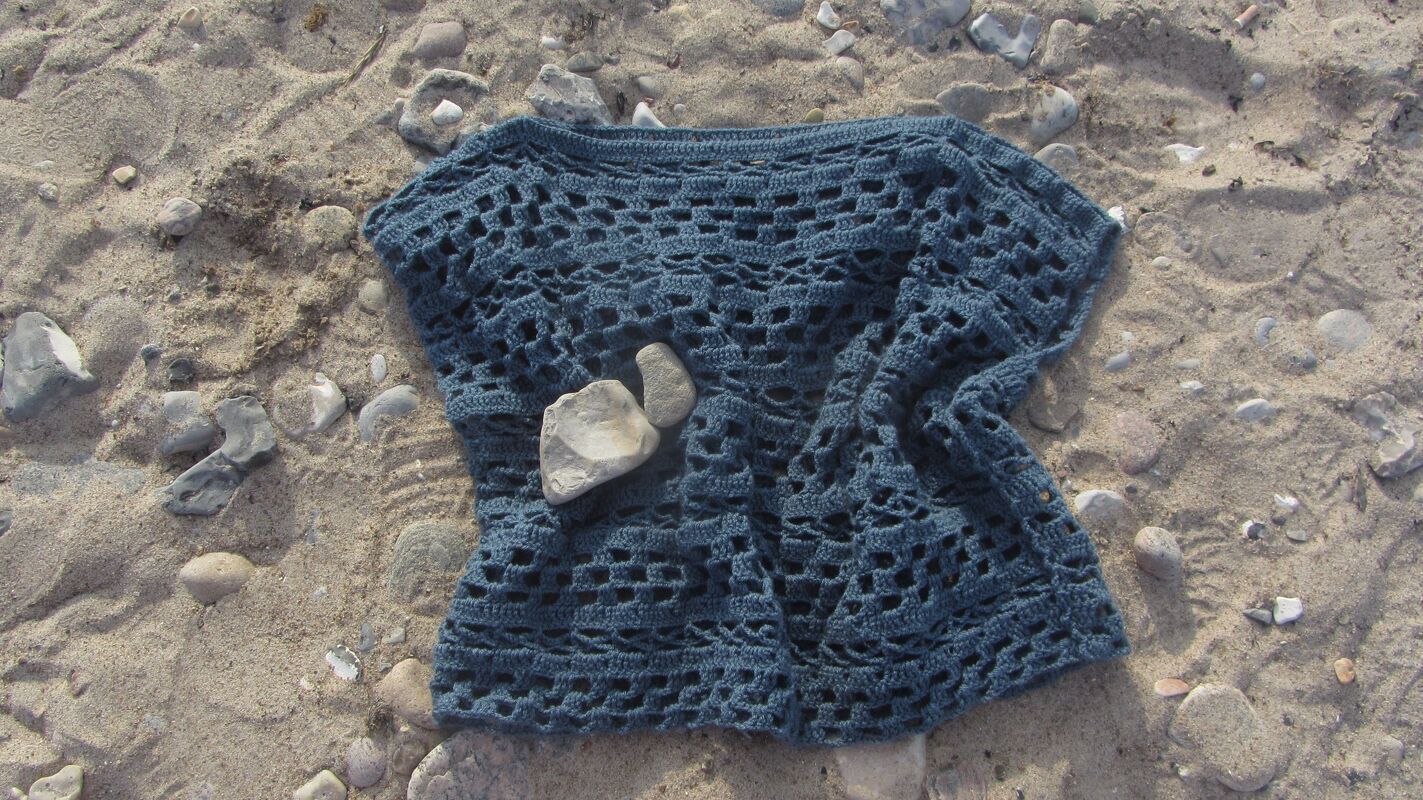 Dunkelblaues Damen-Häkelshirt mit Lochmuster auf sandigem Boden und Kieselsteinen