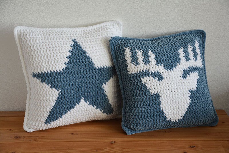 Zwei gehäkelte Kissen mit Stern- und Elchmotiv auf Holzfläche