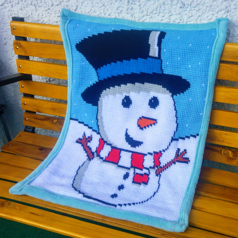 Gehäkelte hellblaue Babydecke mit Schneemann, Zylinder und gestreiftem Schal auf einer Holzbank