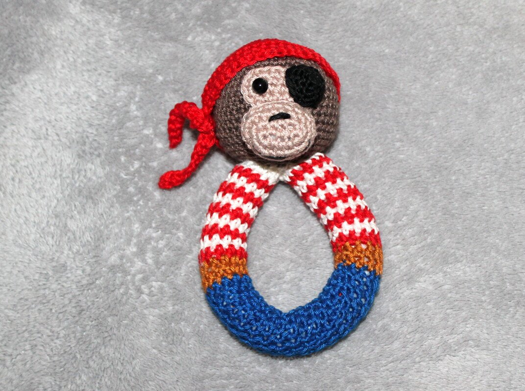 Crocheted pirate monkey rattle with red bandana, eye patch and striped ring on a grey background.