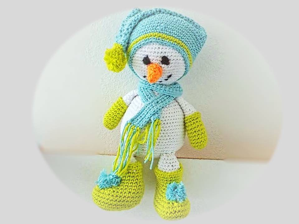 Gehäkelte Schneemann-Puppe in Weiß mit hellblauer Mütze, Schal sowie grünen Fäustlingen und Stiefeln.