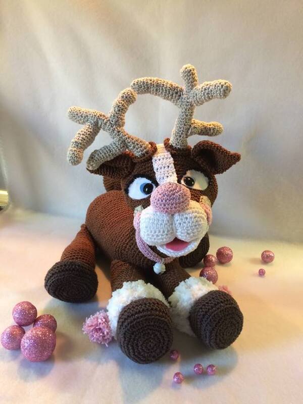 Gehäkeltes braunes Rentier-Amigurumi mit rosa Nase, weißer Schnauze und beigen Geweihen, umgeben von rosa Glitzerkugeln.