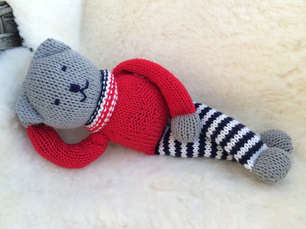 Gestrickter grauer Teddy mit rotem Pullover und blau-weiß gestreiften Beinen, liegend auf hellem Fell