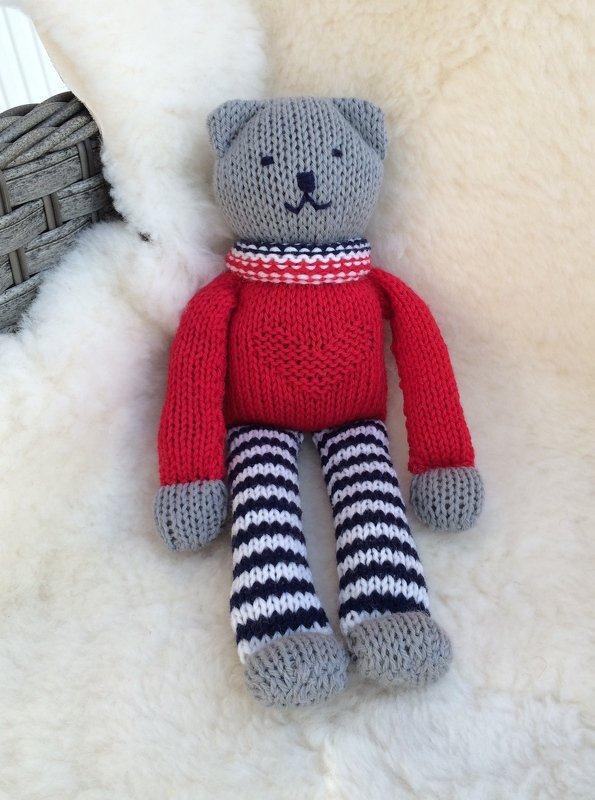Gestrickter grauer Teddybär mit rotem Herzpullover und schwarz-weiß gestreiften Beinen auf hellem Fell