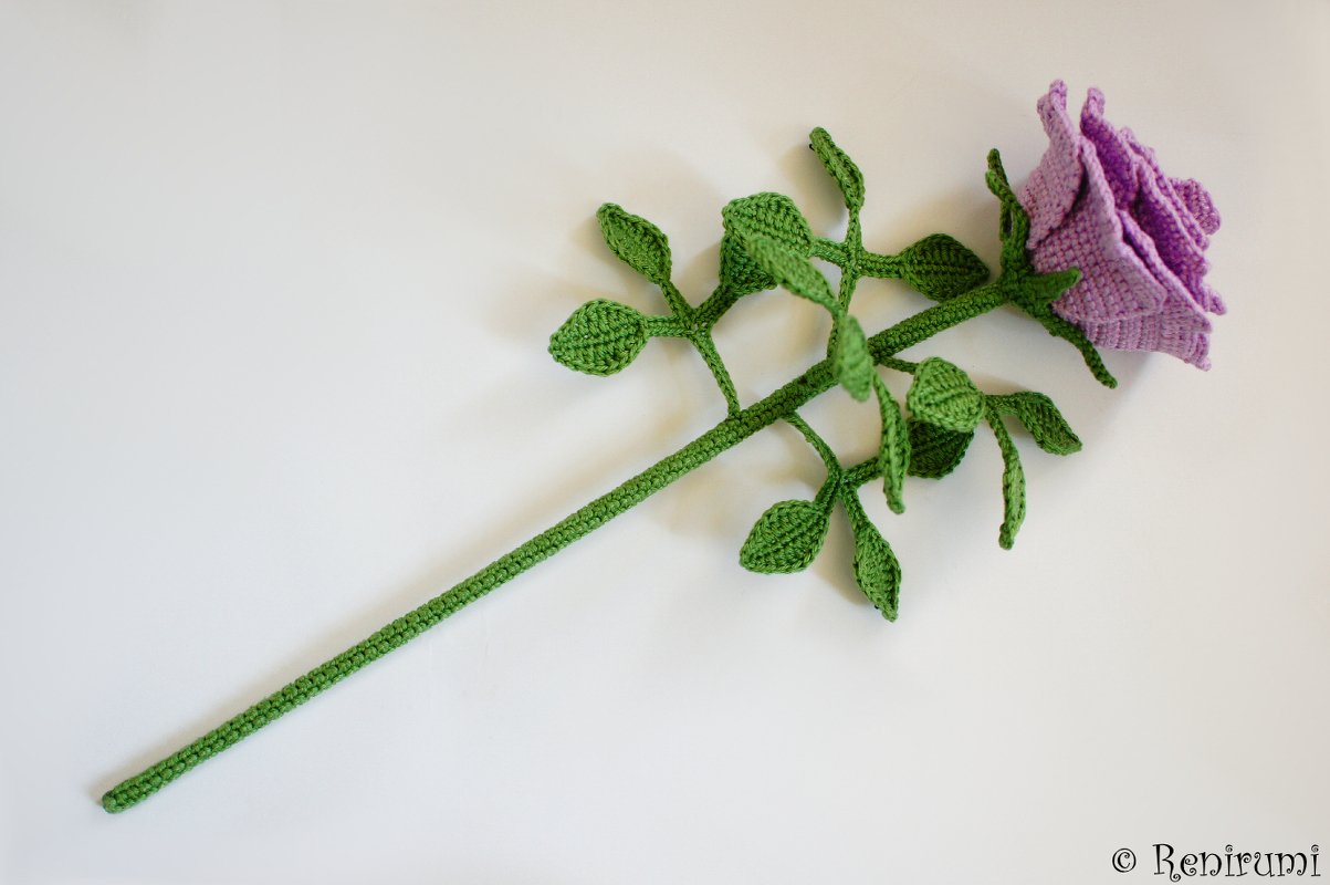 Purple crocheted rose with a long green stem and leaves on a white background.