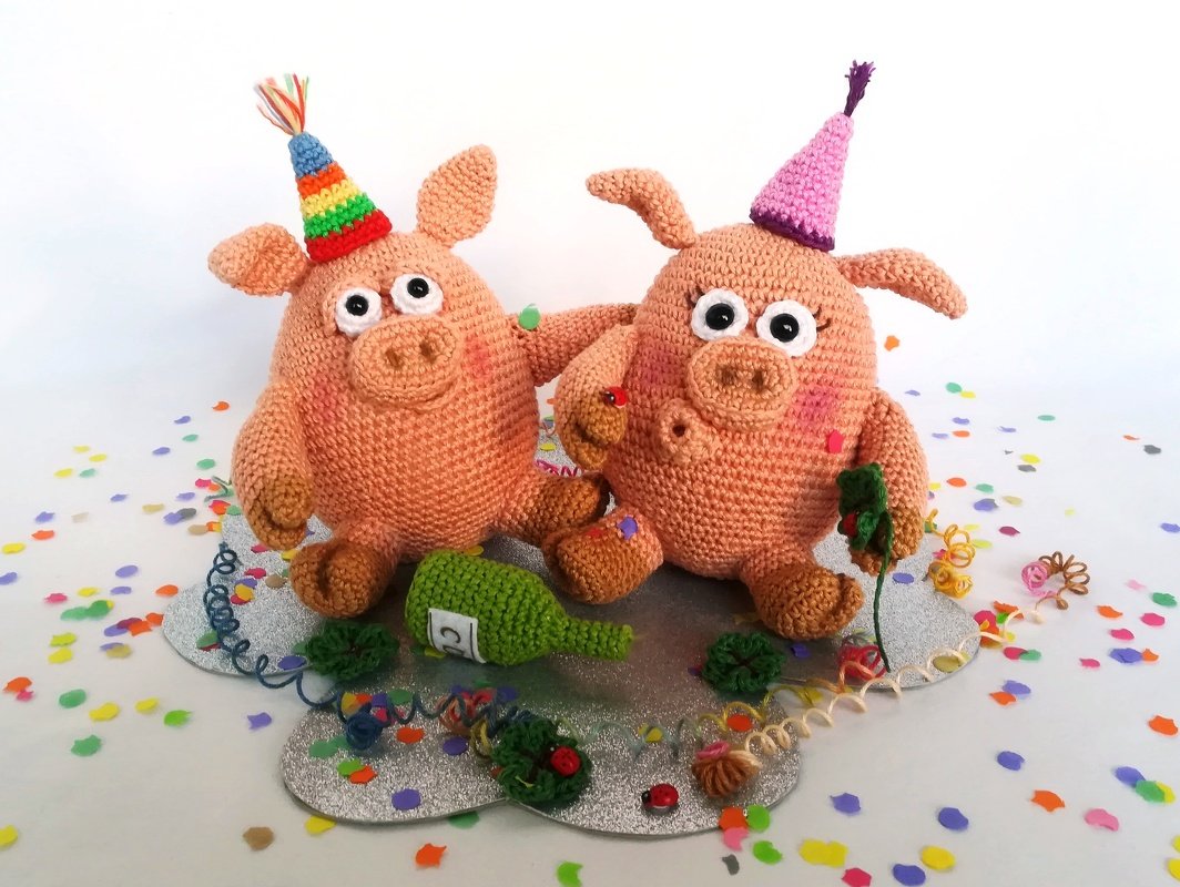 Two crocheted pig amigurumi wearing colorful party hats seated on confetti with a small green crocheted bottle