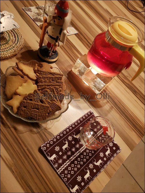 Gepolstertes Mug-Rug mit Rentiermuster, Glas-Teekanne und Teller mit Weihnachtsplätzchen auf Holztisch