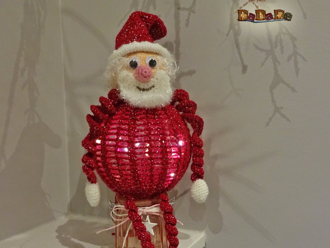 Gehäkelter Weihnachtsmann mit rotem Glitzerkörper und weißem Bart, sitzend auf einem Glasgefäß.