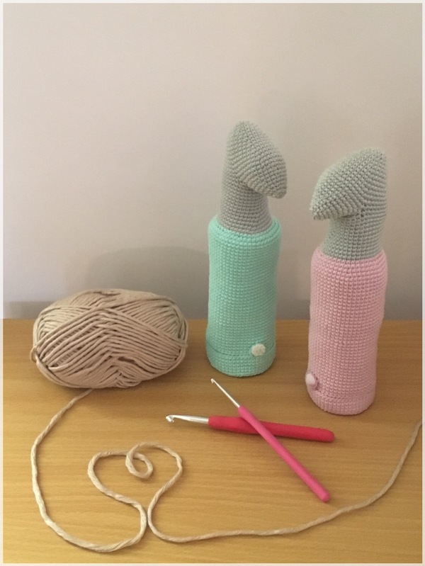 Zwei zylindrische gehäkelte Utensilos in Mint und Rosa mit Garnknäuel und Häkelnadeln auf einer Tischfläche