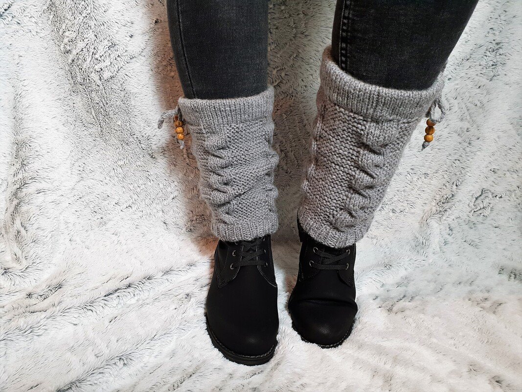 Beine in Jeans und schwarzen Stiefeln mit grauen gestrickten Beinstulpen, Zopfmuster und Kordel mit Holzperlen