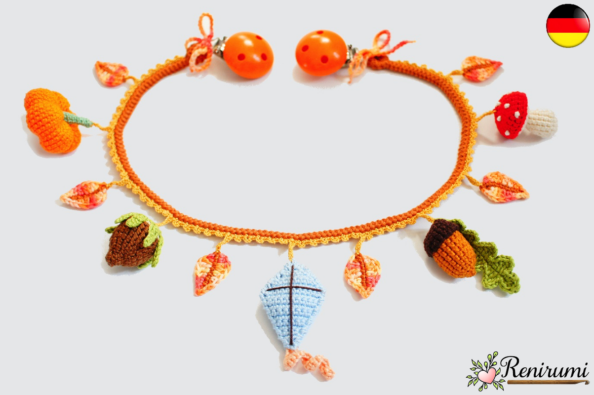 Gehäkelte Herbstgirlande mit Kürbis, Fliegenpilz, Eicheln, Tannenzapfen, bunten Blättern und blauem Drachen-Anhänger auf hellem Hintergrund.